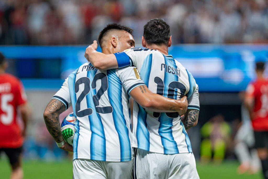 Argentina players Lautaro Martinez and Lionel Messi embracing on the pitch in their blue and white striped kits after a goal, with Egypt players in red in the background