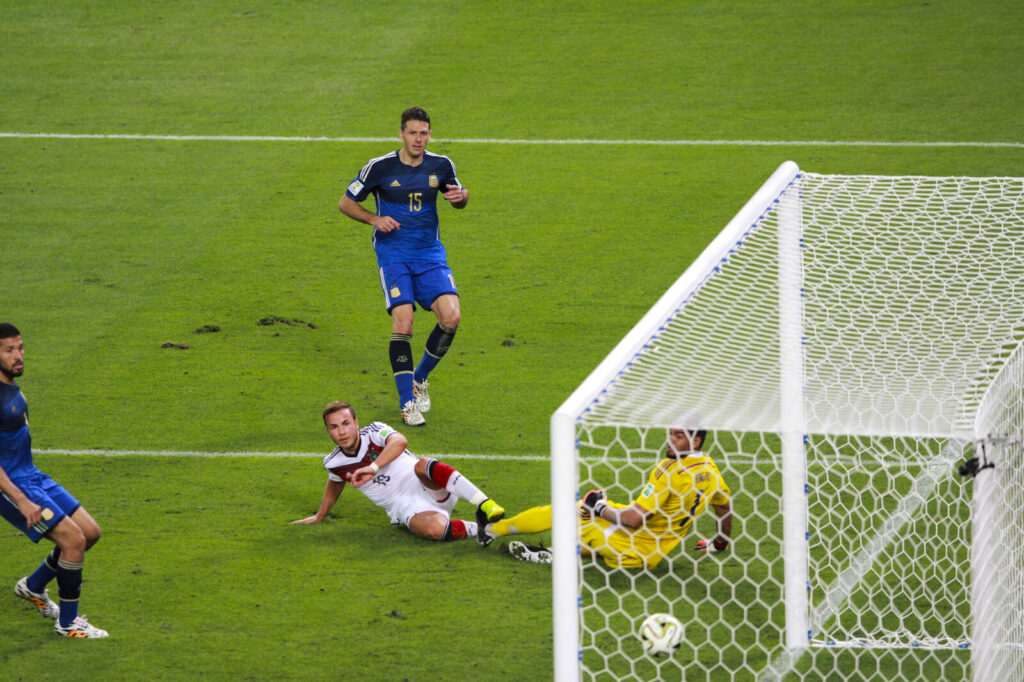 Soccer player striking the ball to shoot on goal during a match, with a defender and goalkeeper in the frame