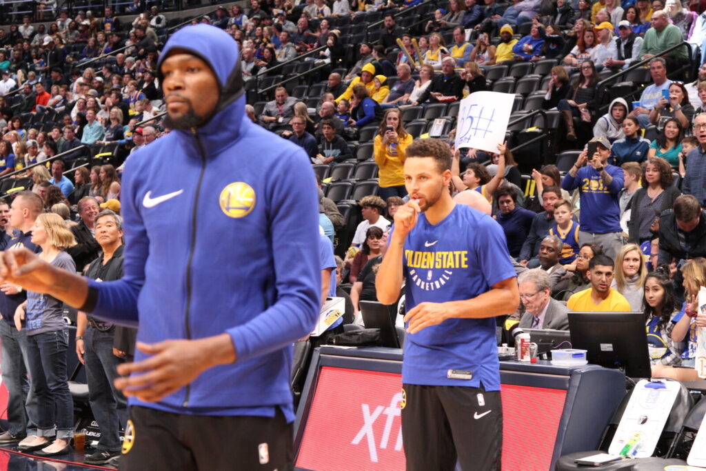 Golden State Warriors players Kevin Durant and Stephen Curry stand courtside in team gear as fans in Warriors jerseys fill the stands behind them.