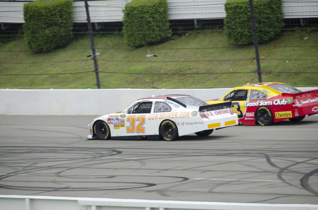 Two NASCAR stock cars racing side by side on an oval track, with tire marks visible on the asphalt.