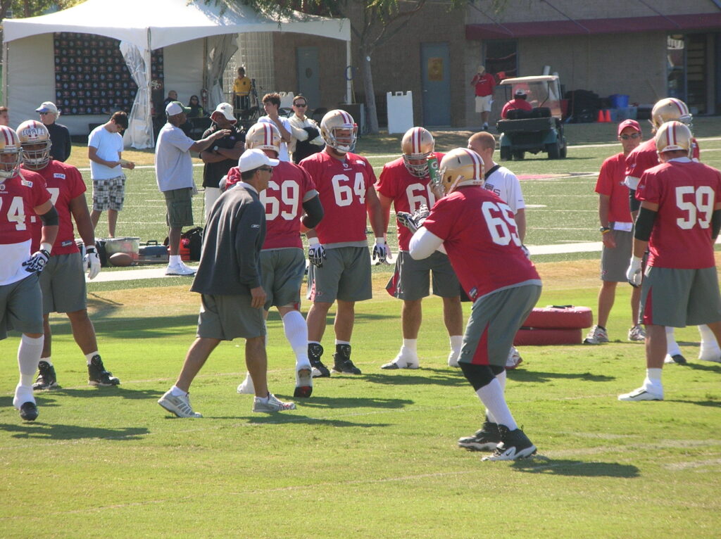 San Francisco 49ers offensive linemen huddle with a coach in full practice gear during 2010 training camp.