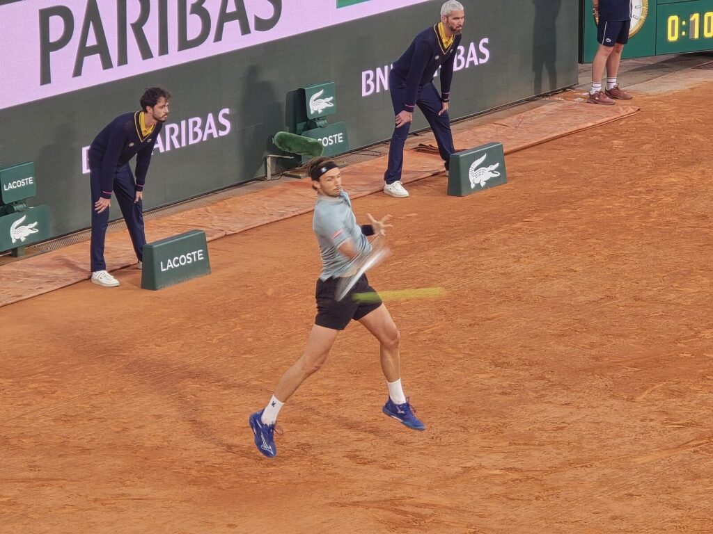 Jannik Sinner in mid-swing forehand on a clay court at Roland Garros, ball boys and BNP Paribas/Lacoste boards visible in the background. (Foto de arquivo: Jannik Sinner em Roland Garros 2025, não em Monte Carlo 2026.)