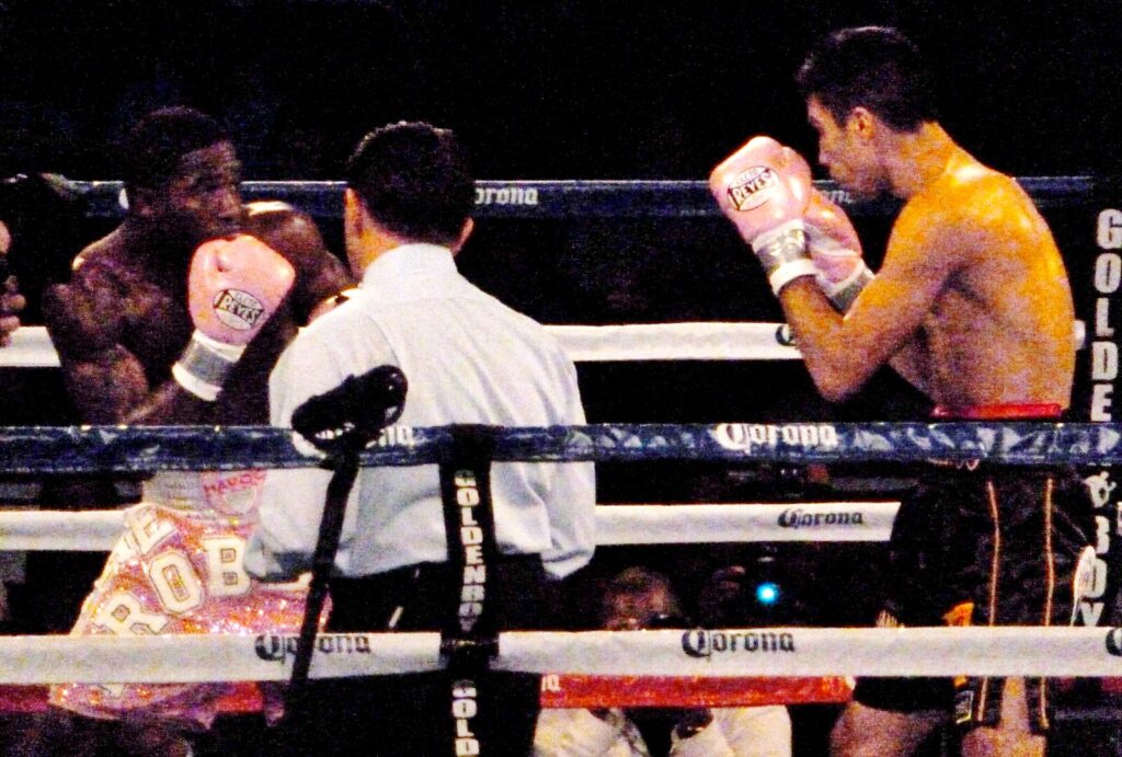 Two boxers exchange punches during a professional bout, with a judge scoring the action ringside.