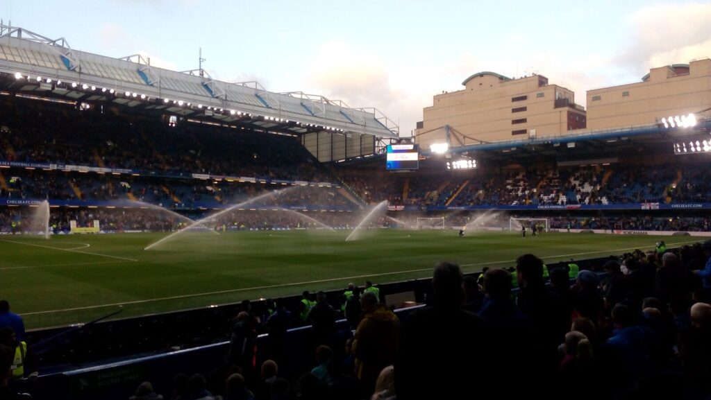 Wide view of Stamford Bridge stadium during a match, pitch sprinklers running, fans filling the stands