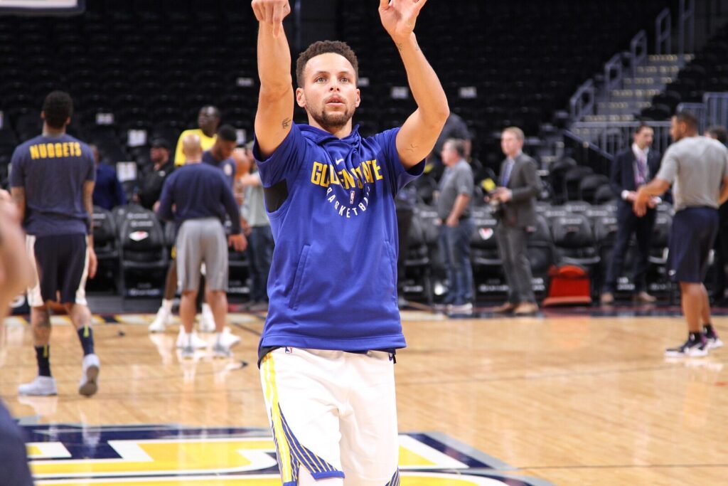 Stephen Curry in a Golden State Warriors hoodie and shorts, warming up on court pregame with teammates and staff visible in the background at an away arena (File photo: Stephen Curry warming up before a Warriors road game.)