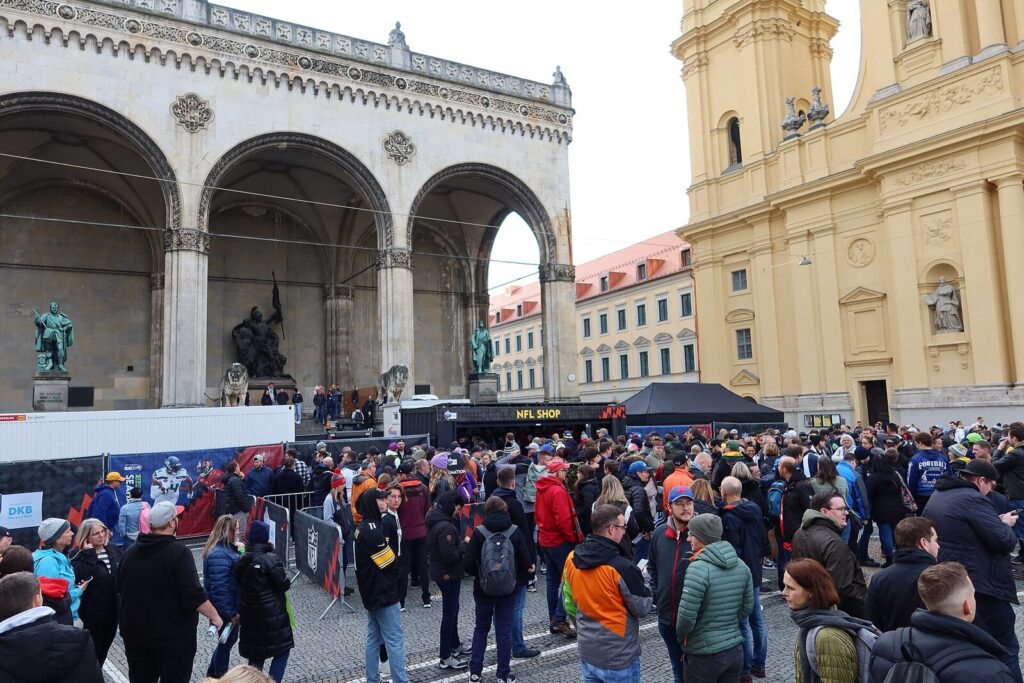 Crowd at an NFL fan shop plaza outside a historic building, with a partially visible Seattle Seahawks banner among NFL team promotional material (Foto de arquivo: público em evento externo da NFL em Munique, 2022, sem relação direta com a abertura da temporada 2026.)