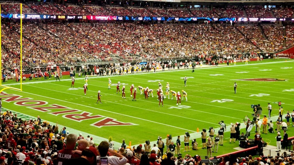 Wide-angle stadium shot of an NFL game between the Packers and Cardinals at State Farm Stadium, October 2025, with players on the field and a full crowd