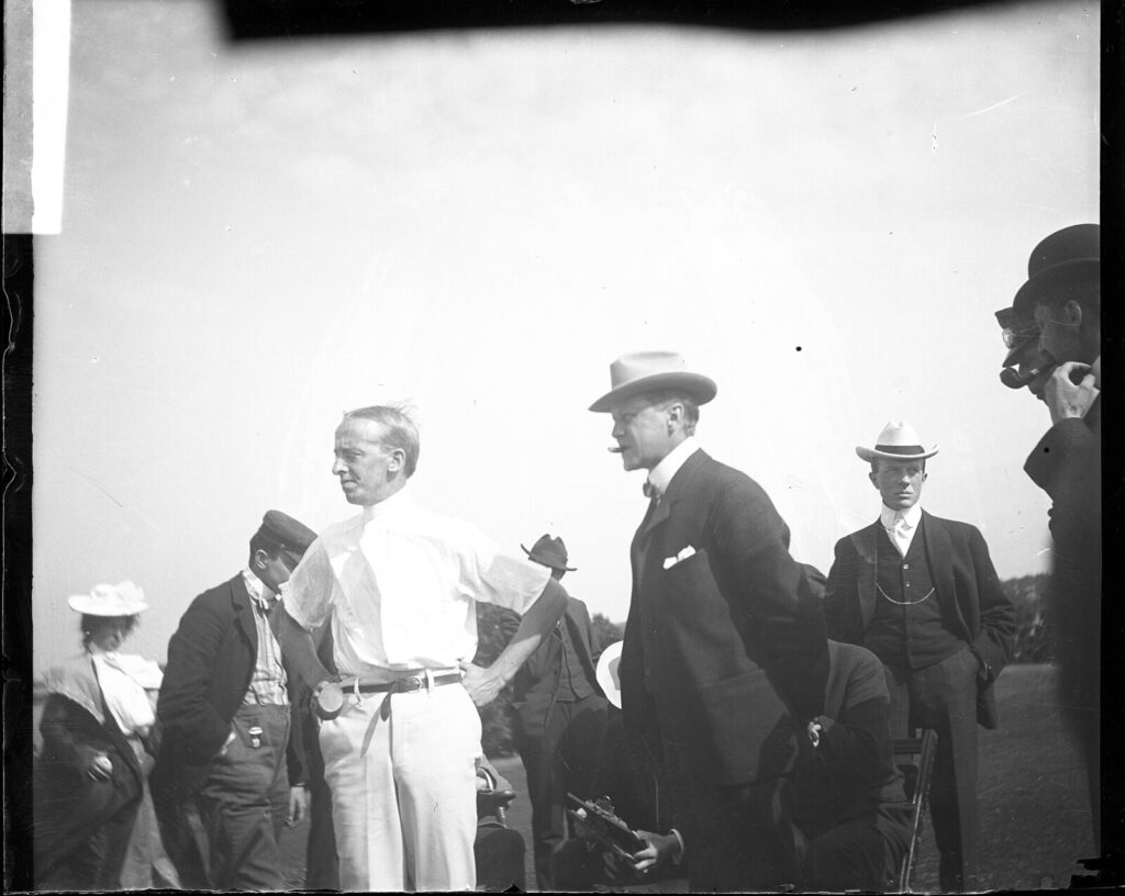 Black-and-white early sideline photograph of an American football game, players and a coach standing near the field with a packed grandstand behind them (Historical file photo of an early American football sideline (University of Minnesota), used for generic illustration and unrelated to any specific current NFL practice-squad transaction.)